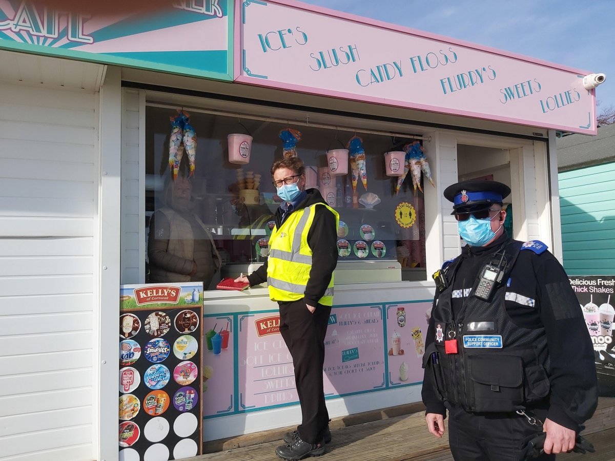 Male in hi-vis jacket and facemask is talking to a kiosk owner at the seafront across the counter. He is accompanied by a police community support officer wearing a face-mask.